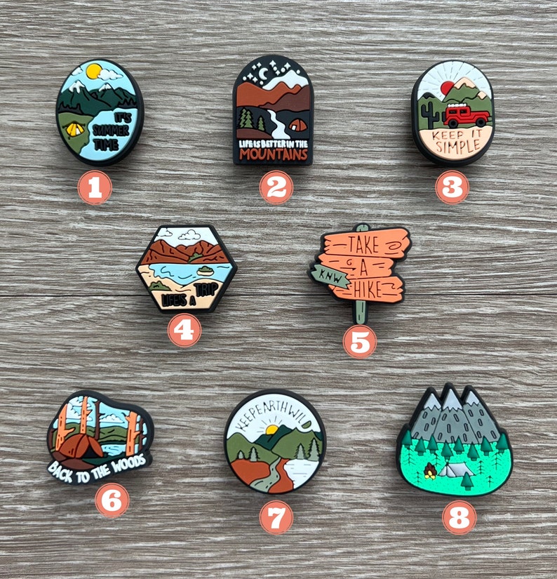 May include: A collection of eight camping-themed shoe charms. Each charm features a different design, including mountains, tents, and phrases like "Keep It Simple" and "Life is Better in the Mountains." The charms are in various shapes and colors.