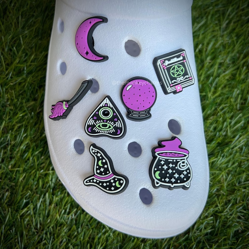 Op de afbeelding: Een witte schoen versierd met heksenthema-bedels. Bedels zijn onder meer een halve maan, een bezem, een Ouija-bord, een kristallen bol, een spreukenboek, een heksenhoed en een ketel. De bedels zijn zwart, paars en groen.