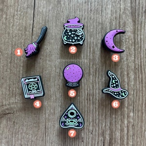 Op de afbeelding: Een verzameling zwarte en paarse Halloween-charms. De charms omvatten een bezem, ketel, halve maan, spreukenboek, kristallen bol, heksenhoed en een planchette. De charms zijn gerangschikt op een houten oppervlak.