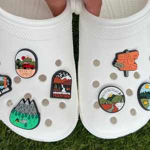 May include: White clogs adorned with colorful, nature-themed charms. The charms feature designs like mountains, forests, a red truck, and text that reads "Keep It Simple" and "Take a Hike." The charms are in various shapes and sizes.