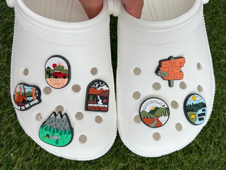 May include: White clogs adorned with colorful, nature-themed charms. The charms feature designs like mountains, forests, a red truck, and text that reads "Keep It Simple" and "Take a Hike." The charms are in various shapes and sizes.