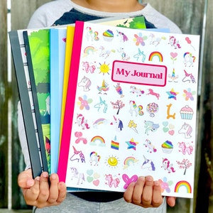 May include: A child holds a stack of colorful notebooks. The top notebook is white with a pink border and features a unicorn theme with the text "My Journal".