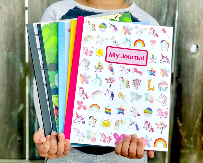 May include: A child holds a stack of colorful notebooks. The top notebook is white with a pink border and features a unicorn theme with the text "My Journal".