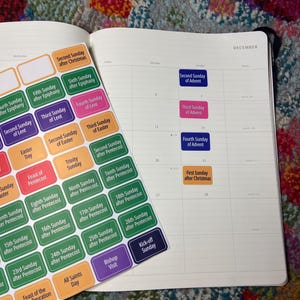 May include: An open planner with a December calendar page and a sheet of colorful stickers. The stickers have text like "Second Sunday of Advent" and "Third Sunday of Lent". The planner is on a patterned surface.