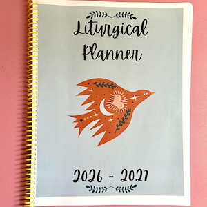 May include: A spiral-bound liturgical planner with a light blue cover featuring a central orange bird illustration decorated with a white crescent moon, a heart, and stars. The text reads Liturgical Planner and 2026-2027 in black script.