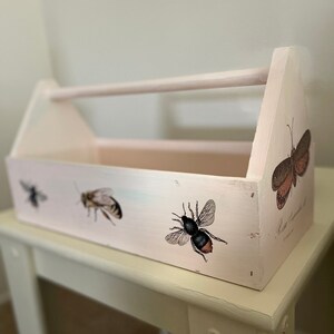 May include: A white wooden toolbox with a handle, featuring a vintage-inspired design with bee and butterfly illustrations. The toolbox has a distressed paint finish and is perfect for organizing small items.