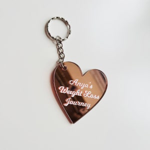 May include: Rose gold heart-shaped keyring with a silver key ring. The keyring has the text "Anya's Weight Loss Journey" engraved in white.