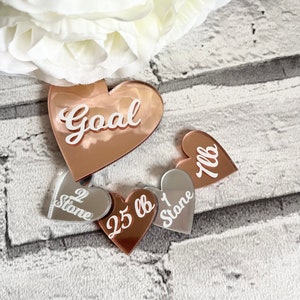 May include: Five rose gold and silver heart-shaped weight loss motivational plaques. The largest plaque reads "Goal". The other plaques read "1 lb", "2 stone", "25 lb", and "1 stone".