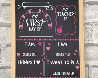 May include: A framed chalkboard with a light wood border. The black chalkboard has pink and white text that reads "My First Day Of" and "My Teacher Is". It includes fill-in-the-blank sections for age, height, and aspirations.