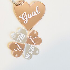 May include: A set of four rose gold and clear acrylic heart-shaped keychains. The keychains are connected together to form a clover shape. The top keychain has the word "Goal" written on it. The other three keychains have the words "Stone", "7lb", and "6'2" written on them.