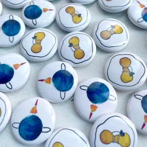 May include: A collection of white, round buttons with hand-drawn illustrations. Some buttons feature a blue and orange design, while others display a yellow hourglass-like figure with a black outline. The buttons are scattered across a light surface.