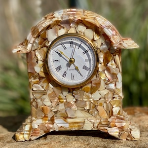 May include: A small, decorative clock made of seashells. The clock face is white with black Roman numerals and gold hands. The clock is sitting on a wooden surface.