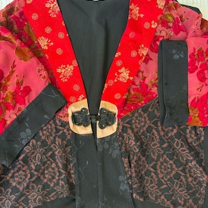 May include: A black jacket featuring red panels with gold floral patterns and circular symbols. The garment includes black lace overlays on the lower sections, black cuffs, and a central beige fabric closure with black knotted frog fasteners.