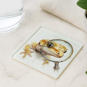 May include: Square glass coaster featuring a watercolor illustration of a gecko, viewed through a magnifying glass. The gecko has yellow and brown markings. The magnifying glass has a silver handle.