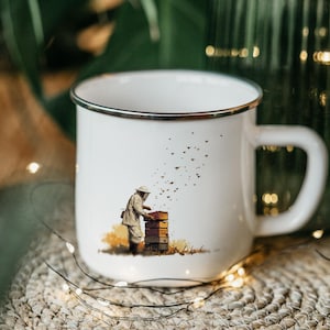 May include: White enamel mug with a silver rim featuring a beekeeper tending to a beehive, with bees flying around. The mug has a large handle and is set against a blurred background with greenery and fairy lights.