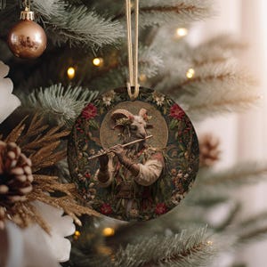 May include: Round Christmas ornament featuring a goat-like figure playing a flute, surrounded by flowers and foliage. The ornament has a gold string for hanging. The background is a Christmas tree with lights and other ornaments.