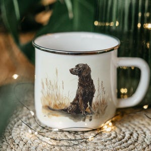May include: White enamel mug with a black rim and handle. The mug features a detailed illustration of a brown dog sitting in tall grass. The background is blurred, with a green plant and string lights.
