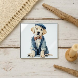 May include: Square glass coaster featuring a watercolor illustration of a Labrador puppy dressed in a blue blazer, bow tie, and cap. The puppy is sitting on a white background. The coaster is surrounded by a wooden background.