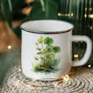 May include: White enamel mug with a silver rim and handle. The mug features a watercolor illustration of aquatic plants and small fish. The design includes green leaves and a red and silver fish. The mug is on a woven mat.