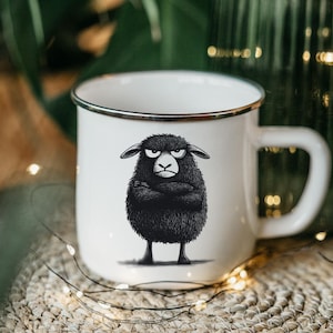 May include: White enamel mug with a black rim and a black and white illustration of a sheep with a grumpy expression. The sheep is standing with its arms crossed.