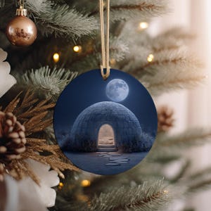 May include: Round Christmas ornament featuring a nighttime scene with an igloo under a full moon. The igloo has a lit doorway, and the background is a dark blue sky. The ornament is hanging from a gold string.