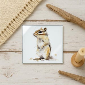May include: Square glass coaster featuring a watercolor painting of a chipmunk. The chipmunk is depicted in shades of brown, tan, and white, with a detailed rendering of its fur and features. The coaster is set against a white background.