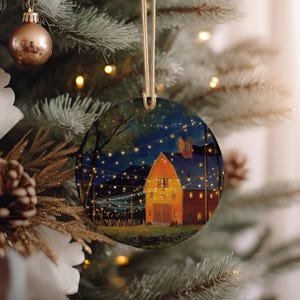 May include: Round Christmas ornament featuring a nighttime scene of a barn illuminated by string lights. The ornament has a gold string for hanging and is displayed on a Christmas tree with other ornaments and lights.