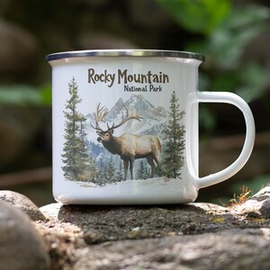 May include: White enamel mug featuring a majestic elk in a mountain landscape, with the text "Rocky Mountain National Park." The mug has a silver rim and a white handle. The design includes pine trees and mountains.