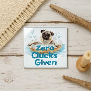 May include: Square glass coaster featuring a pug dog floating on a donut in a pool of water. The coaster has the text "Zero Clucks Given" in teal. The background includes small blue birds.