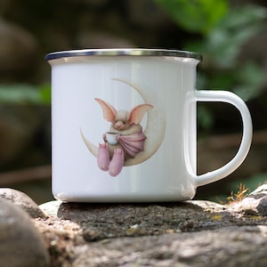 May include: White enamel mug with a silver rim, featuring a whimsical illustration of a bat-like creature seated on a crescent moon, holding a cup. The creature is depicted in soft pink and beige tones. The mug has a white handle.