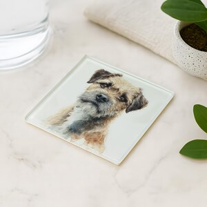 May include: Square glass coaster featuring a watercolor portrait of a Border Terrier dog. The coaster has a white background and the dog is depicted in shades of brown, black, and white. The coaster is on a white surface.
