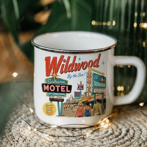 A white mug with a vintage-style illustration of a motel called Wildwood by the Sea, featuring neon signs and people walking on a street.