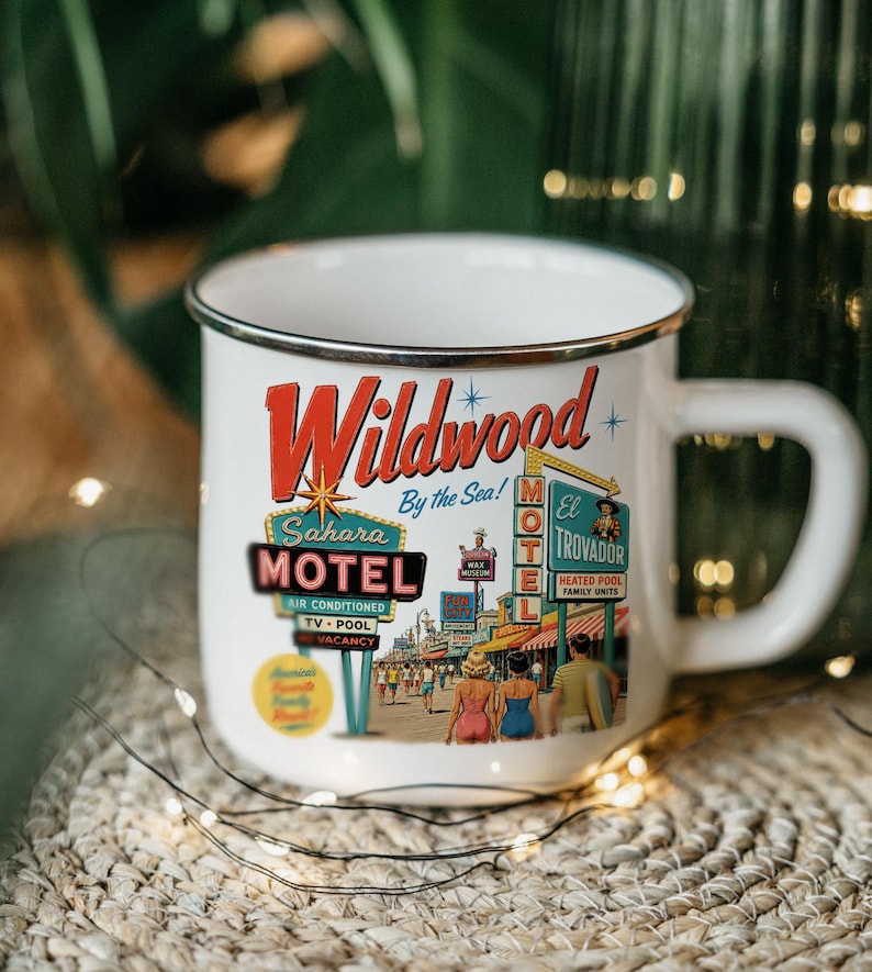 A white mug with a vintage-style illustration of a motel called Wildwood by the Sea, featuring neon signs and people walking on a street.