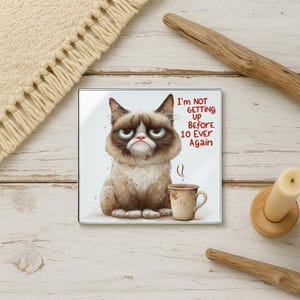 May include: Square glass coaster featuring a grumpy cat illustration with the text "I'm NOT GETTING UP BEFORE 10 EVER AGAIN." The cat is brown and white, with a mug of coffee. The coaster is on a light wood surface.
