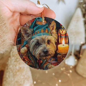 May include: Round Christmas ornament featuring a dog wearing a blue hat and colorful coat. The dog is in front of a lit candle and a toy soldier. The ornament has a gold string for hanging.