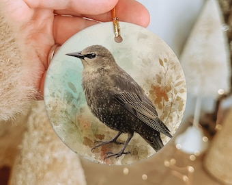 May include: Round ceramic ornament featuring a detailed watercolor illustration of a brown bird with a black beak and legs. The bird is set against a soft, muted background with floral accents. A gold string is attached for hanging.