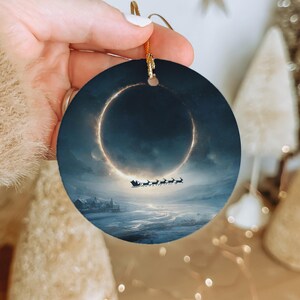 May include: Round Christmas ornament featuring a winter landscape with Santa's sleigh and reindeer silhouetted against a celestial ring. The ornament has a gold string for hanging and is held by a hand.