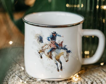 May include: White enamel mug with a silver rim and a handle. The mug features a watercolor painting of a cowboy riding a bucking bronco. The cowboy is wearing a blue shirt and hat. The horse is white.