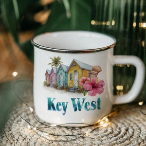 May include: White enamel mug with a black rim, featuring a watercolor illustration of colorful beach houses, a palm tree, and a pink hibiscus flower. The words "Key West" are printed in blue. The mug has a large handle.