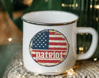 May include: White enamel mug with a black rim and handle. The mug features a distressed circular design of the American flag with the word "patriot" in red letters. The mug is sitting on a woven surface.