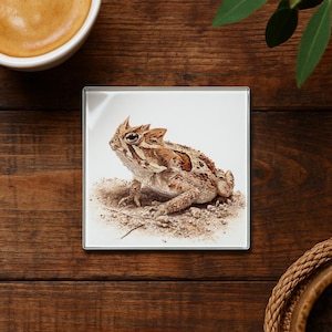 May include: Square coaster featuring a detailed illustration of a horned lizard. The lizard is depicted in shades of brown and tan, with a textured appearance. The coaster has a glossy finish and a silver edge, suitable for home decor.