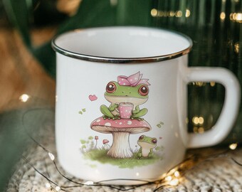 May include: White enamel mug with a silver rim featuring a green frog wearing a pink bow sitting on a pink and white mushroom with a smaller green frog sitting on the mushroom.