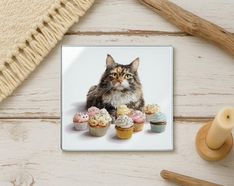 May include: A square glass coaster featuring a fluffy cat surrounded by cupcakes with colorful frosting. The cat has brown, black, and white fur and is looking directly at the viewer. The cupcakes have pink, yellow, and blue frosting.