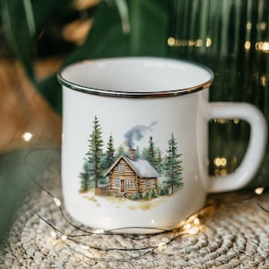 May include: White enamel mug with a silver rim featuring a watercolor illustration of a log cabin in the woods. The cabin has a brown roof and a chimney with smoke coming out of it.