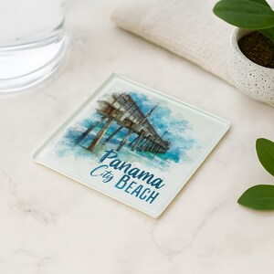 May include: Square glass coaster featuring a watercolor image of a pier over turquoise water. The coaster has the words "Panama City Beach" in blue script. A glass of water, a small potted plant, and a sprig of leaves are in the background.