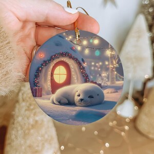 May include: A round Christmas ornament featuring a sleeping white seal pup in a snowy winter scene. The scene includes a snow-covered house with a red door, string lights, and a small gift box. The ornament has a gold string for hanging.