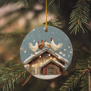 May include: Round Christmas ornament featuring a winter scene with chickens on a snowy coop roof. The ornament has a light blue background with a string of lights and is hanging from a gold string.