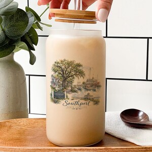 a hand opening a glass jar with a printed design of a coastal scene, including a tree, boats, and a building. The jar is placed on a wooden surface, with a wooden spoon nearby. There is also a bottle and a vase in the background, adding to the overall composition of the scene.