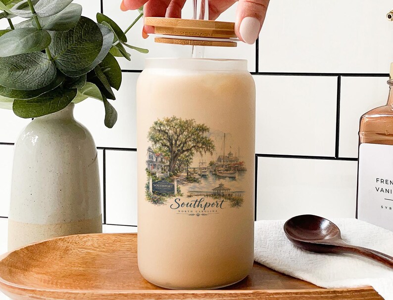 a hand opening a glass jar with a printed design of a coastal scene, including a tree, boats, and a building. The jar is placed on a wooden surface, with a wooden spoon nearby. There is also a bottle and a vase in the background, adding to the overall composition of the scene.