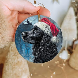 May include: Round Christmas ornament featuring a black dog wearing a Santa hat. The dog's portrait is set against a blue background with a painting of a person fishing. The ornament has a gold string for hanging.